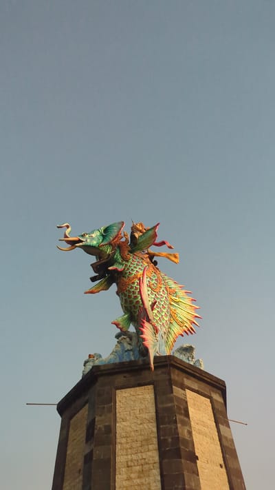 The image shows a colorful, ornate dragon-fish sculpture attached to a stone structure with a blue-gray sky in the background. The sculpture features green, orange, and red colors and intricate scale and fin details. The stone structure appears to be part of a building or temple, and is located in Pererenan, Indonesia.