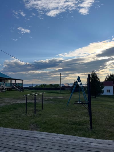 This media file depicts an outdoor scene in Krasnoturansk, Russia, likely during the late afternoon or early evening given the warm, golden light illuminating the clouds. The sky is a prominent feature, with dramatic, streaky clouds catching the sun, creating a sense of depth and brilliance. The overall weather appears to be fair, with a clear blue sky dominating the upper portion of the image and scattered white clouds drifting by.
The scene includes a grassy, somewhat unkempt field with a dirt path winding through it. Visible objects include a children's swing set with blue A-frame supports and black horizontal bars. Further back, there are parallel bars, suggesting a small outdoor fitness area. A small, simple white house with a red roof and a single window is nestled amongst trees in the background. Another structure, possibly a small shed or an annex to a house, is visible with a green, slatted exterior. A wooden deck or platform occupies the right foreground, constructed from weathered wooden planks laid horizontally.
The image is taken from a slightly elevated perspective, looking out over the field towards a body of water in the distance, beyond which are rolling hills or mountains. A thin power line stretches diagonally across the frame from the bottom left towards the sky. There are no people visible in the image, and the overall mood is peaceful and serene, characteristic of a rural or semi-rural setting in the evening. No text is visible in the media file.