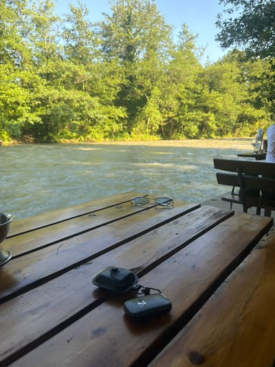 The image captures a serene outdoor scene at a riverside cafe. In the foreground, a weathered wooden table holds a pair of sunglasses and two black key fobs. The table is positioned to overlook a moderately flowing river, its surface rippling under natural light. Lush green trees line the opposite bank of the river, creating a vibrant backdrop against a clear blue sky. In the background, several outdoor seating areas with wooden benches and tables are visible, suggesting a public space for dining or relaxation. The overall atmosphere is peaceful and natural, indicative of a tranquil setting likely located in Abzhanda, Abkhazia. There are no people clearly visible in the frame.