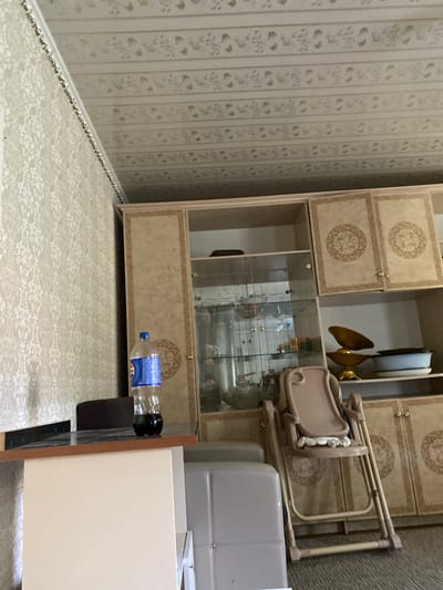 The image shows an indoor scene, likely a living room or dining area. A large, ornate cabinet with glass doors and decorative panels dominates the right side of the frame. On a counter in front of the cabinet, a partially filled bottle of Pepsi is visible. To the left of the counter, a laptop is present. In the foreground, a beige baby high chair is folded and leaning against the cabinet. The walls are covered in patterned wallpaper, and the ceiling features a decorative embossed pattern. The flooring appears to be carpeted. The overall impression is of a domestic setting in Миң-Булак, Kyrgyzstan.