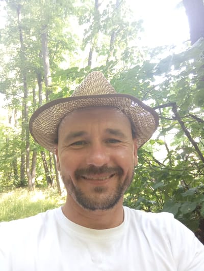A man is taking a selfie outdoors, in a wooded area with trees and foliage. He is wearing a woven straw hat and a white t-shirt. The man has a beard and is smiling at the camera. The sun is bright, suggesting it is daytime. The background shows a blur of green leaves and trees, with some light filtering through the branches. There are no visible signs of text in the image.