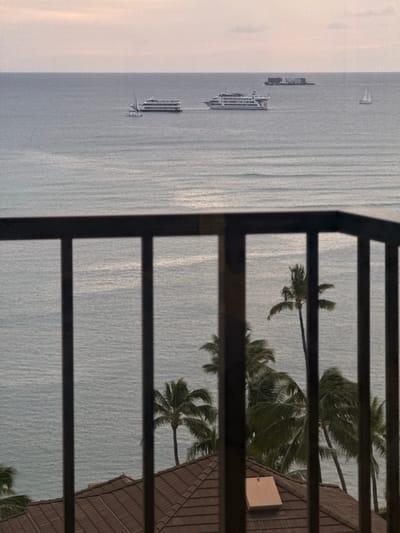 The image shows a view from what appears to be a balcony, likely in Honolulu, United States of America. The foreground is dominated by a dark, vertically-oriented railing. Through the railing, the scene unfolds: the ocean takes up a significant portion, with several ships and a sailboat visible on the left side of the frame. Additionally, palm trees are scattered, interspersed with a glimpse of rooftops in the vicinity.