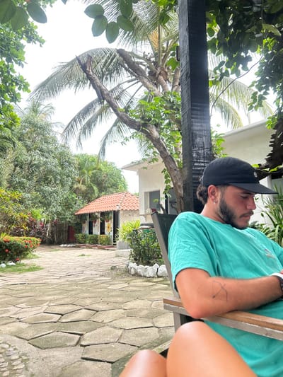 A young man with a black baseball cap is sitting on a chair in a courtyard, looking down. He is wearing a light teal t-shirt and has a tattoo on his left forearm. The chair is positioned in front of a thick, dark wooden pole. His legs are visible, and he appears to be relaxed. The courtyard is paved with gray stone slabs and lined with bushes and palm trees. A small building with a red tile roof is visible to the left of the image. The man is in the shade of a large palm tree and other tropical vegetation, which surrounds the courtyard. The location, according to the coordinates, is on the beachfront of Palomino, Colombia.