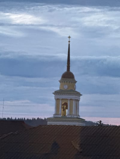 A church bell tower stands against a cloudy sky, topped with a cross. The tower is yellow with white trim and features a clock face and visible bell. Below the tower, a dark, tiled roof occupies the foreground. A line of trees is visible in the middle ground, and a hint of pink light suggests either dawn or dusk. The bell tower is a prominent landmark in the city of Selizharovo, Russia.