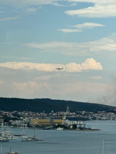 The image shows a panoramic view of the Seget Donji area in Croatia. In the upper portion of the frame, an airplane is visible in the sky amidst fluffy white clouds against a light blue sky. Below the sky, a dark green, tree-covered hillside forms a backdrop to the scene. Along the coastline, a city with white buildings and red-tiled roofs stretches out. In the foreground, a body of water, likely the Adriatic Sea, is dotted with numerous sailboats and yachts moored in a harbor. A large industrial building with a yellow facade and a construction crane are also visible on the waterfront, suggesting a harbor or shipyard. The overall scene depicts a coastal town with maritime activity and an airplane in the sky.