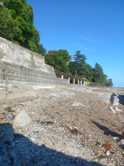 The image depicts a partially constructed or dilapidated concrete structure on a rocky shoreline under a clear blue sky, suggesting a sunny day. The structure appears to be a terraced or stepped embankment made of rough concrete and stone. Green trees and foliage are visible on the upper slopes behind the structure, suggesting a natural, possibly coastal, setting. The ground in the foreground and on the structure itself is covered with small stones and gravel. There's a plastic bottle with a blue cap visible on the ground near the lower left of the image, which is a common litter item. The lighting and shadows indicate it is likely midday. The scene evokes a sense of abandonment or a work-in-progress, with no people visible. The context provided, Gwandra, Abkhazia, places this scene in a specific geographical region known for its coastline and historical sites.