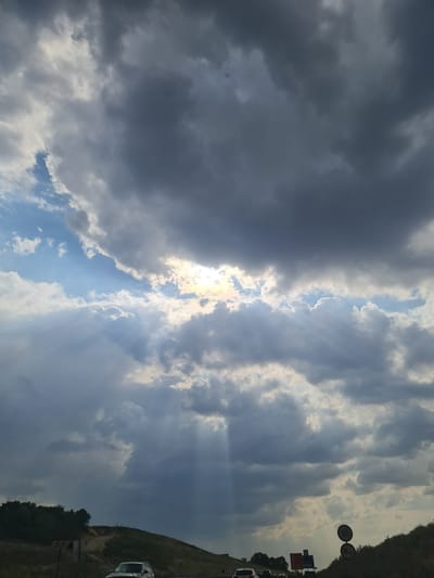 The image captures a dramatic sky with heavy, dark clouds interspersed with beams of sunlight breaking through. The sun is partially visible behind a break in the clouds, casting rays downwards. Below the sky, a road is visible, with cars driving on it, suggesting it is a highway. The landscape on either side of the road includes grassy hills and some vegetation. Road signs are also present, including a speed limit sign showing "55". The overall impression is of a dynamic weather event over a transportation route, possibly on the outskirts of a city. While the image does not explicitly show the city of Tatabánya, Hungary, the presence of a speed limit sign in kilometers per hour (55) is consistent with Hungarian road regulations, and the terrain with hills is also characteristic of the region around Tatabánya, known for its mining landscape and hilly terrain.