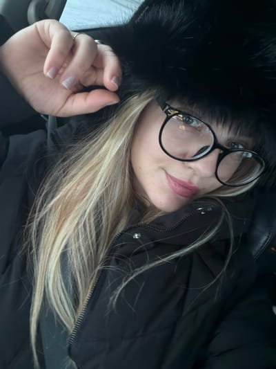 A female subject with blonde hair is shown in a close-up, chest-up orientation, seated. She is wearing a black, padded winter jacket and a black fur-trimmed hat or hood. Black-framed, round eyeglasses are present, along with pink lipstick. Her left hand, displaying a silver ring on the ring finger and a light-colored manicure, is positioned near the top of her headwear. The subject maintains direct eye contact with the viewer, exhibiting a subtle smile. The blurred background suggests an indoor environment, likely within a vehicle, with an indistinct light-toned area visible in the upper left. The scene is situated in Азьмушкино, Russia.