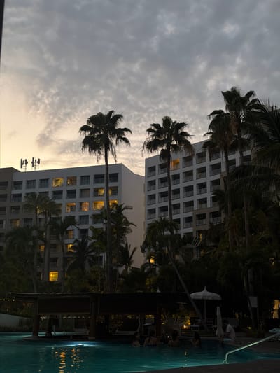 The image shows a building in Las Jarretaderas, Mexico, near a swimming pool. Palm trees are visible in the foreground. The building is multistory with numerous windows, some illuminated. The sky is overcast. People are using the swimming pool and surrounding area. The pool is reflecting light, indicating it is nighttime or dusk.