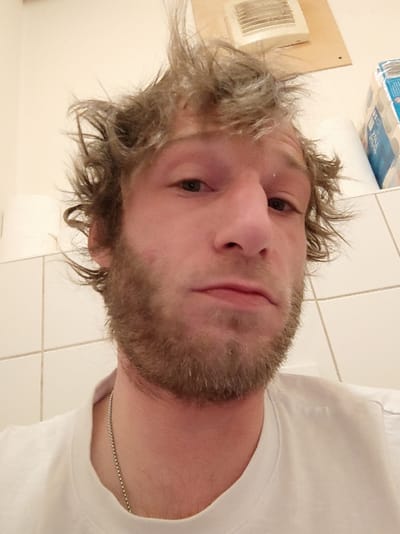 A close-up photograph depicts a male subject in an interior setting, identified as being in Constance, Germany. The individual has light skin, disheveled light brown hair, and a light brown beard, with slight erythema noted on the viewer's left cheek. He is wearing a white crew-neck t-shirt and a thin, silver-colored ball chain necklace. His gaze is directed forward. The background features light-colored square wall tiles. In the upper right portion of the image, a white exhaust fan is mounted on a light-colored wooden panel, and a stack of paper rolls, including one with blue and white packaging, is visible on a surface. A plain, light-colored wall is present in the upper left background.
