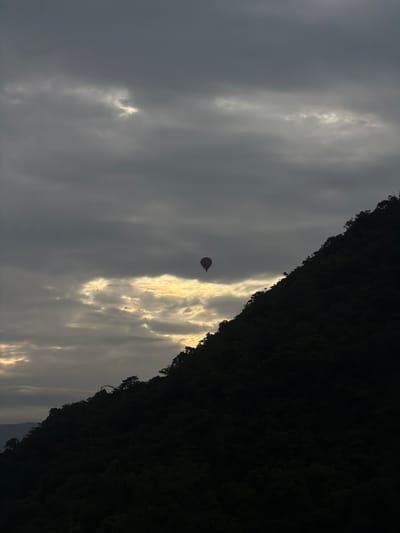 A hot air balloon, silhouetted in red, floats in a cloudy sky above a dark, tree-covered mountain slope. Sunlight breaks through the clouds in a golden band behind the balloon. The image appears to have been taken in Hong Ngern Village, Laos, during twilight or a similarly overcast period.