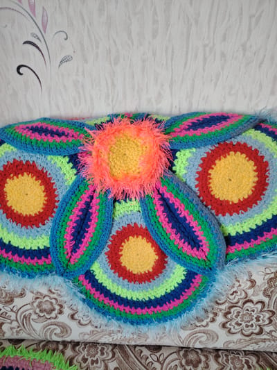 The image displays a vibrant, multi-colored crocheted item, likely a decorative pillow or a section of a blanket, resting on a patterned couch against a textured wall. The crocheted piece is the focal point, featuring a large, central flower-like design. Its core is made of fluffy, neon orange yarn, creating a striking textural contrast. Surrounding this center are petal-like or concentric circular patterns, meticulously crocheted in bright rings of yellow, red, lime green, various shades of blue (including sky blue and royal blue), and hot pink. The distinct stitches highlight the handmade nature of the craft. Some edges of the crocheted item are finished with a light blue, fluffy yarn border.
Behind the textile, a light-colored wall with subtle patterning is visible. The wallpaper features delicate, swirling vine or floral motifs in shades of grey, black, and soft pinkish-purple, appearing to have a slightly textured or shimmery finish. Below the crocheted item, a portion of a couch or sofa is upholstered in a light-colored fabric patterned with intricate brown and beige floral and swirling designs. Another section of similar patterned fabric is partially visible beneath, suggesting a layered or folded textile.
No activity is depicted in the static scene. The lighting is bright and even, characteristic of an indoor setting, and does not indicate a specific time of day. There is no visible text within the image. The overall impression is one of vibrant, handcrafted decor in a domestic environment in Yanaul, Russia.