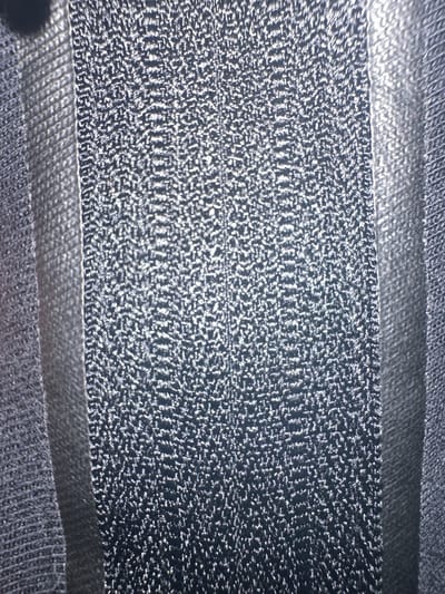 This is a close-up, abstract image of a dark, textured material, possibly fabric or a woven surface. The dominant colors are dark gray and black, with bright white highlights scattered across the surface, suggesting a rough or slightly reflective texture. The material appears to be layered, with distinct horizontal bands. One band, in the center, has a more intricate, granular texture with a shimmering quality. Above and below this central band are smoother, darker bands. The lighting creates strong contrasts, with bright reflections on the textured areas and deep shadows elsewhere. There is no discernible scene, setting, or identifiable objects. No people are visible, and no activity or event is taking place. There is no visible text in the image. The location context of Letterkenny, Ireland, is not visually represented in the image itself.