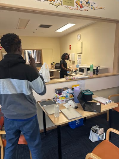 This image captures an indoor scene, likely a breakroom or common area, in Carrum Downs, Australia. In the foreground, a person with dark, curly hair wearing a black and gray hoodie and blue jeans stands facing away from the camera, holding a mobile phone.

In the midground, a light-colored wooden L-shaped counter or table is filled with various items. These include an open black laptop, a closed gray laptop, a clipboard with papers, multiple water bottles (one purple, one clear), snack items like "K-TIME" bars and "M&M's," paper napkins, and keys. An "ALDI" branded blue shopping bag rests on the floor next to the table. Orange chairs with metal frames are partially visible around the table.

Behind the counter, a woman with dark, curly hair, dressed in a dark short-sleeved top and pants, is standing. She appears to be engaged in an activity within a kitchenette area, which includes a microwave and other appliances. The background wall features a clock, a hand towel dispenser, and a red emergency device. Overhead, multiple light fixtures illuminate the room. The left wall displays decorative floral patterns and partially visible text that reads "COMMUNITY," "UNDS," and "DRADIO FONDS." The floor is covered with dark carpet.

The overall scene suggests a casual environment, possibly a community center or workplace, with individuals taking a break or attending to tasks. The time of day is not discernible due to artificial lighting.