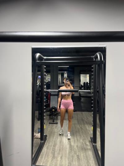 The image shows a woman taking a selfie in a gym. She is wearing a pink sports bra and pink shorts, and white athletic shoes. She has headphones on and is holding a smartphone in her right hand. The gym appears to be modern, with grey wood-like flooring and black gym equipment. In the background, through the reflection in the mirror, other gym equipment and some posters on the wall are visible. The lighting suggests it is daytime, but no specific time is discernible. The scene is typical of a workout session at a fitness facility. No discernible text is legible in the image or its reflection. The user's query specifies "Altötting, Germany" as a location context, which is not visually confirmed by the image itself.