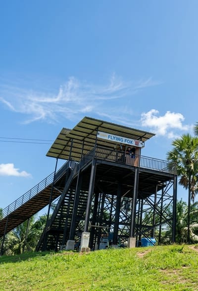 This is a low-angle, full shot of a metal, multi-level structure with a covered top platform, located in a grassy, outdoor area surrounded by palm trees under a bright blue sky with sparse white clouds. The structure has a long, inclined ramp leading to its lower level and a steep staircase to the upper level. A sign on the upper platform reads "FLYING FOX" with a logo. Two people are visible on the upper platform, standing and looking out. At the base of the structure, there are several concrete supports, informational signs, and a blue barrel. The overall scene suggests an adventure park or recreational facility in Bintang Meriah, Indonesia, likely a starting point for a zip-line activity.