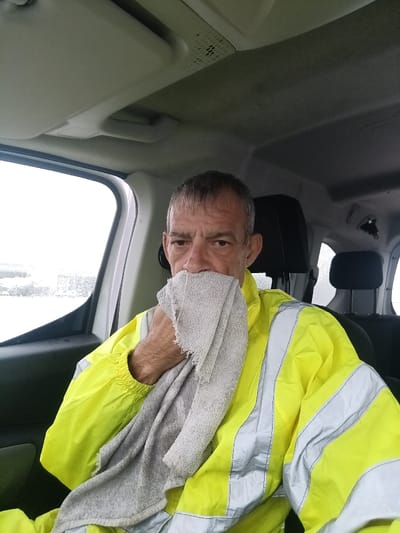 An adult male is seated inside a vehicle in Ojos de Garza, Spain. The subject, appearing middle-aged with short, greying hair, directly faces the camera. He is wearing a bright yellow, long-sleeved, high-visibility jacket with horizontal and vertical silver-grey reflective stripes. His right hand holds a light grey, textured, and possibly frayed towel, positioning it over his mouth and nose. The vehicle's interior exhibits a light-colored headliner with an overhead console and a sun visor. A dark headrest is visible behind the subject. A window to the subject's left reveals a bright, indistinct exterior environment, suggesting overcast weather or low visibility conditions.