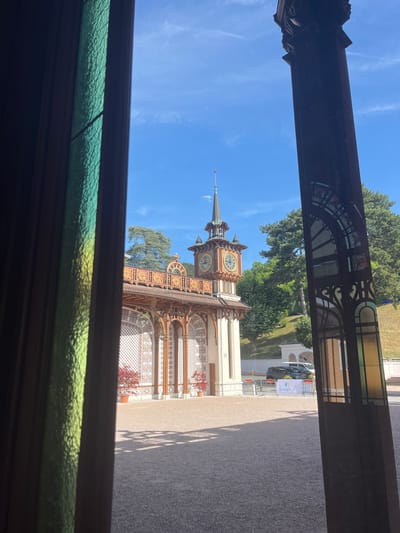 The image depicts a view through a stained-glass window and a dark wooden frame, looking out onto a courtyard. In the center of the courtyard stands a multi-story building with a clock tower. The building has intricate wooden latticework, decorative gold accents, and a tiled roof. Red potted plants are visible in front of the building. The courtyard is paved with gravel. A banner with the "Evian" logo is visible in the background, along with some vehicles and safety barriers. Lush green trees and a sloped grassy hill are situated behind the building. The sky is a clear, bright blue with a few wispy clouds. The scene appears to be from Évian-les-Bains, France, likely near the Thermes Évian, given the presence of the Evian branding and the architectural style. No people are clearly visible in the image.