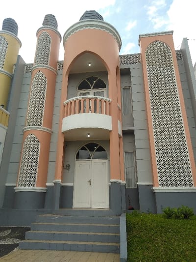 This image depicts a multi-story building located in the city of Nathene, Laos, viewed from a low angle under an overcast sky. The building's facade is characterized by a peach-colored central cylindrical tower featuring an arched recess on the second floor, which contains a white balustraded balcony and a pair of white-framed arched-top doors. A single light fixture is visible within this arch. The ground floor beneath the balcony presents the main entrance with white double doors, each featuring a single horizontal handle, topped by an arched transom window.
Flanking the central tower are two decorative, peach-colored, minaret-like structures, each integrated into the building's light gray walls and capped with a dark gray, ridged roof. These structures incorporate tall, vertical panels of intricate white geometric latticework. The main wall sections are light gray, with a contrasting dark gray band along the base and outlining architectural elements. A horizontal frieze with repeating geometric patterns adorns the upper section of the building. Vertical, rectangular windows with white frames are visible on the side walls; a dark-handled broom or similar tool leans against the wall below the rightmost window.
Access to the entrance is provided by a series of gray concrete steps. The ground in front of the steps is paved with light-colored pavers. To the left, a section of dark and light gray patterned paving is visible, while to the right, a sloped patch of green grass with small, round-leafed plants is present. No people or discernible actions are depicted.
