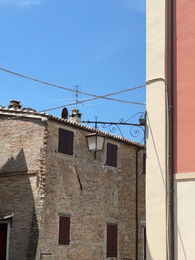 The image depicts an external view of an old brick building in Barbara, Italy. The building has a tiled roof and several dark brown shutters on its windows. A vintage-style lantern hangs from an ornate wrought-iron bracket attached to a pole. The building's brickwork shows signs of age and weathering.
In the foreground and right side of the image, a section of another building is visible, painted in two distinct colors: a light cream color and a pinkish-orange color. A dark downspout runs vertically along the cream-colored section, with several clips securing it to the wall.
The sky above is a clear, bright blue. Several wires crisscross the sky, and an antenna is mounted on the roof of the brick building. The overall scene suggests a sunny day in a historic Italian town.