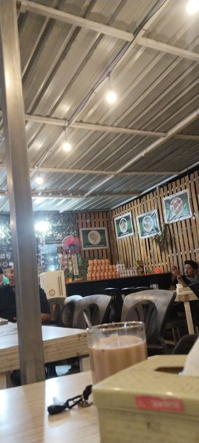 The image depicts an indoor dining area, likely a cafe or restaurant, in Peurupok, Indonesia. The ceiling is constructed with corrugated metal sheets supported by metal beams, and several circular light fixtures are visible. The walls are adorned with wooden planks and several framed pictures or posters, some featuring circular graphics with text. To the left, a large silver cylindrical pillar stands prominently. In the background, a counter area is visible with a water dispenser, stacks of canned beverages, and various items arranged on shelves. Seating includes chairs and tables, with some patrons present. In the foreground, a glass of beverage and a tissue box are placed on a table. One person is visible on the left side of the image, partially obscured by the pillar. Another person is seated on the right, looking at a phone. The lighting suggests it is evening or indoors with artificial light.