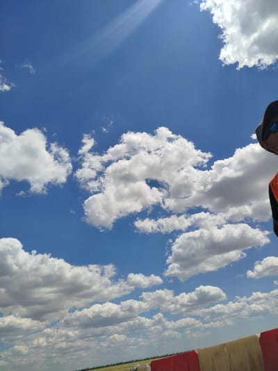 A clear blue sky with scattered cumulus clouds is visible. In the foreground, there is a portion of a red and white barrier. To the far right, a person wearing a dark cap and sunglasses is partially visible, with an orange vest or shirt. The landscape below the sky appears to be a field with a treeline in the distance. The location is Komyshuvate, Ukraine.