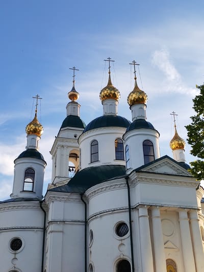 The image shows a white orthodox church with golden domes against a blue sky with some clouds. The church has multiple domes, some of which are tall and slender, while others are bulbous. Each dome is topped with a cross. The church has arched windows and classical architectural elements like columns. A bell tower is visible in the background with bells hanging inside. The church is surrounded by greenery, with the leaves of a tree visible on the right side of the image. The sun appears to be setting or rising, casting a warm light on the building. The location is identified as the city of Chelganovo, Russia. There are no people visible in the image.