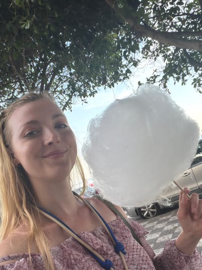 The image is a selfie taken from a low angle, likely by a woman with blonde hair. She is holding a large piece of white cotton candy on a stick. She appears to be standing on a sidewalk with trees and a parked car visible in the background. The location is likely Chianchitta, Italy.