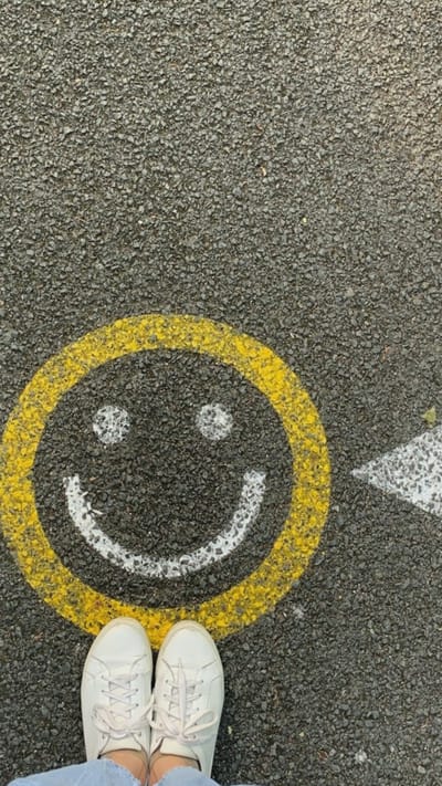 The image shows a close-up, overhead view of a smiley face painted on asphalt pavement. The smiley face is outlined in yellow and features white dots for eyes and a white curved line for a mouth. Two white sneakers are visible at the bottom of the frame, indicating someone is standing over the smiley face. A white arrow pointing to the right is partially visible on the right side of the smiley face. The asphalt is dark gray and textured with small stones. The overall scene is a playful and simple street art or playground marking. The location is Oreshki, Russia.