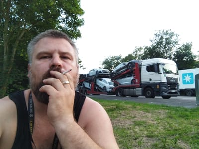 A man in a black tank top is taking a selfie outdoors, holding a lit cigarette to his mouth. He has grey hair, a beard, and is wearing a ring on his right hand. In the background, a car carrier truck is parked, loaded with multiple white cars. To the right of the car carrier is a white trailer with a blue star logo. The scene appears to be along a road with grass and trees. The location is the A27 Autobahn near Loxstedt, Germany.