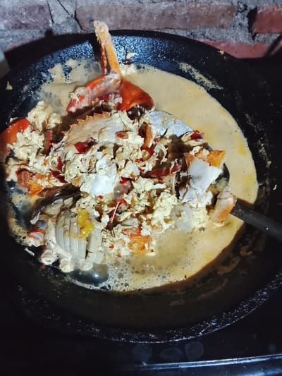 A close-up, overhead shot captures a dish of crab being cooked in a black wok. The wok is filled with a creamy, yellow sauce that appears to be simmering, with small bubbles visible on the surface. Pieces of crab, including claws and shell fragments, are mixed into the sauce along with what looks like scrambled egg and possibly some chili flakes. A silver serving spoon is partially submerged in the wok, resting on the crab and sauce. The background shows a rustic brick wall, suggesting an outdoor or traditional kitchen setting. The image is likely taken in Gunung Malang, Indonesia, given the context provided.