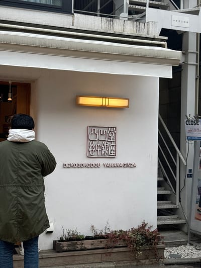 The image shows a white building with a red sign that reads "DEKOBOKodou YANAKAGINZA" with Japanese characters in the center. Above the sign is a rectangular light fixture with an illuminated yellow light. A man wearing a green jacket, blue jeans, and a white hoodie is facing the building with his back turned towards the viewer. He is standing in front of a doorway. The building is located in Arakawa City, Japan at the coordinates 35.728 latitude and 139.766 longitude. There is a sign above the man with Japanese characters on it. There are also stairs leading to the upper level of the building. The background shows a portion of a wooden fence and some foliage.