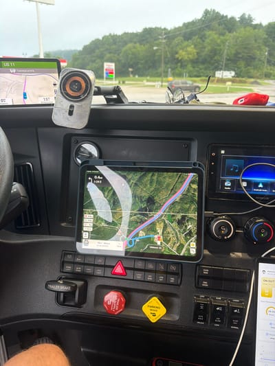 The image shows the interior of a truck's cabin, likely a semi-trailer truck, focusing on the dashboard area. A tablet mounted on the dashboard displays a GPS navigation screen with a route highlighted, showing the location as "Wildwood, GA." Various buttons and controls for the truck's functions are visible, including "TRAILER BRAKE" and "PARKING BRAKE." A secondary display, possibly a car stereo or infotainment system, is also present. In the background, through the windshield, a gas station with visible signage and trees can be seen, suggesting the truck is parked at a service station. The lighting indicates it is daytime, and the sky appears overcast. No people are clearly visible in the immediate cabin area.