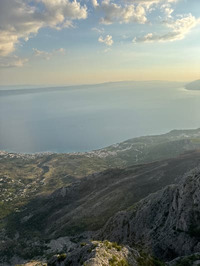 This image depicts a sweeping, elevated view of the coastal area of Općina Tučepi, Croatia. In the foreground, rugged, rocky terrain dominates the lower portion of the frame, with sparse vegetation clinging to the steep slopes. As the view extends towards the middle ground, a verdant, mountainous landscape is visible, characterized by dense green foliage and winding roads that snake across the terrain. A scattering of buildings, indicative of a small settlement, can be seen nestled within the valleys and along the foothills.

The middle ground transitions into a broad expanse of calm, blue water, likely the Adriatic Sea, stretching towards the horizon. A faint line of distant landmasses, appearing as hazy silhouettes, marks the far edge of the sea. The water's surface is disturbed by subtle ripples, and a small, dark shape, possibly a boat, is visible in the distance.

The sky above is a soft gradient of pale blue and warm yellow, with scattered white clouds tinged with sunlight. The overall lighting suggests either early morning or late afternoon, casting long shadows and highlighting the contours of the landscape. The scene conveys a sense of natural beauty and coastal tranquility.