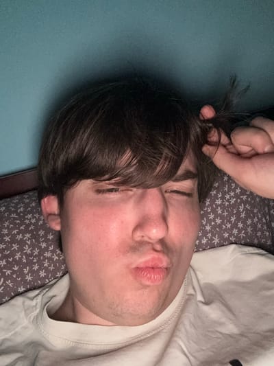 A close-up, low-angle, selfie-style photograph shows a young person with brown hair and fair skin lying down. The person is wearing a white t-shirt and their eyes are closed, with their lips puckered in a kissing expression. Their right hand is raised to their hair, holding a few strands. The person is lying on a pillow with a small white floral pattern on a grey background. The background appears to be a wall painted a teal or blue-green color. The lighting suggests artificial light, possibly from a phone or overhead lamp, creating some shadows and highlights on the person's face and the pillow. The image does not provide any information that would allow for the determination of its location in the city of Dnipro, Ukraine.