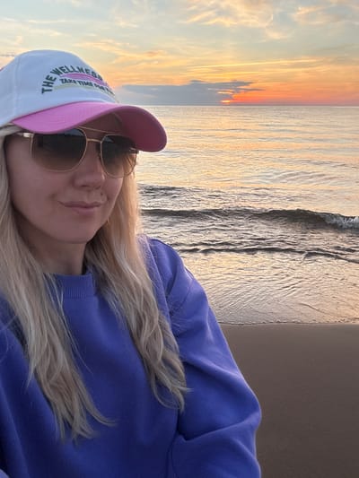A blonde woman, wearing a white baseball cap with pink trim and the words "THE WELLNESS TAKE TIME FOR YOU" in black and pink text, sunglasses with gold frames, and a purple sweatshirt, is standing on a beach at sunset. The ocean is calm with small waves. The sky is filled with colorful clouds in shades of orange, yellow, and pink, indicating the sun is setting over the Baltic Sea. The location is identified as the coast of Kalngale, Latvia. The image is a selfie, with the woman looking towards the camera.