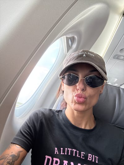 A woman is seated on an airplane, taking a selfie. She is wearing a brown baseball cap with the words "PATRIZIA PEPE" and a small bee logo embroidered on it, and large, dark sunglasses with a geometric frame. Her lips are pursed as if for a kiss, and she has freckles on her face. She is wearing a black t-shirt with pink lettering that reads "A LITTLE BIT DRAMA". Her left arm, visible on the left side of the frame, has tattoos. The airplane's interior is visible, including the textured ceiling, a window, and a gray seat. The image was taken in Hostivice, Czechia.
