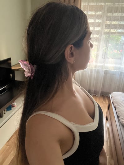 The media file displays a close-up, vertically oriented view of a person's profile, focusing on their head and upper torso. The individual has long, dark hair, partially obscuring their face and neck, and is wearing a black top with white trim. A small, translucent pink hair accessory is visible nestled in the hair. The background suggests an indoor setting with sunlight streaming through a window covered by sheer white curtains, indicating it is daytime. The lighting is soft and diffused. To the right, a wooden surface, possibly part of furniture like a bed or dresser, is visible. The overall impression is one of quiet relaxation or personal reflection. There is no visible text in the media file.