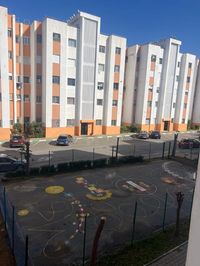 A high-angle view captures a tall, white building with orange accents under a clear blue sky. Parallel to the building runs a road with parked cars. Adjacent to the road is a playground with colorful markings including a hopscotch and various circular designs, bordered by greenery. The composition is oriented vertically, showcasing the building's height and the playground's layout.