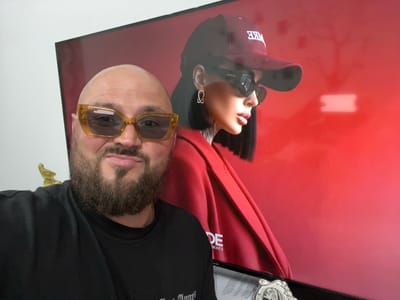 A bald man with a beard and yellow-tinted sunglasses smiles at the camera. He is wearing a black t-shirt. Behind him, a large screen displays a poster of a woman wearing a red jacket, a maroon baseball cap, and sunglasses. The woman has dark hair and is wearing a silver earring. The background of the poster is red. A small golden statue is visible to the left of the man. The man is in the foreground, with the screen behind him filling the rest of the frame. There is no visible indication of the location in Greater London.
