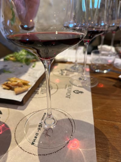A close-up, horizontally oriented shot captures a clear wine glass filled with red wine, resting on a wooden table. The glass is seen from above and slightly to the side, with reflections of light playing across its surface and stem. A human finger is partially visible in the reflection on the glass bowl, suggesting it's being held or recently placed.
The setting appears to be a wine-tasting room or restaurant, likely at a winery in Theewaterskloof Nu, South Africa, as indicated by the location context. In the background, other blurred wine glasses suggest multiple tastings or people. The wine glass is positioned over a paper placemat or menu, which features text and a logo. Some artisanal snacks, possibly cheese or dried fruit alongside green leaves, are visible on a white surface next to the placemat, further reinforcing the wine-tasting or dining context.
Notable details include a red, ring-like light reflection on the paper surface beneath the glass base, likely caused by light refracting through the glass. The text "BEAUMONT" with a logo is visible on the paper placemat. On the base of the wine glass, engraved or printed text reads "Side" and "MOOR-VEH-DRHE" (a phonetic spelling of Mourvèdre). Further descriptive text circles the base, stating, "Mourvedre needs its head in the cool soil & a view of the sea! * The name: an homage to Sebastian's favourite surf spot in One great for hearty foods with to 1974 & 197" with some words cut off, indicating a specific narrative about the wine or winery. The light source creates bright, circular bokeh effects in the background.
