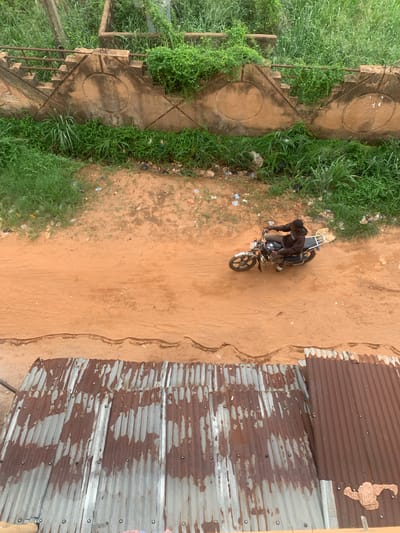 An aerial perspective captures a motorcyclist on a dirt path in Oko Town, Nigeria. The path is bordered on one side by a concrete structure with steps, overrun with greenery. The opposite side features corrugated metal sheets forming a wall. Debris is scattered on the ground adjacent to the path. The motorcyclist is positioned towards the center of the frame.