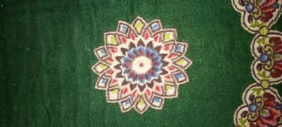 A section of patterned green carpet features a central rosette design. The rosette is multi-colored, with brown, red, blue, and green hues arranged in a concentric pattern. The rosette's shape is radial and symmetrical. To the right, there is a partial view of another patterned element, appearing as a floral border with similar color palettes. The base of the carpet is a solid, textured green.