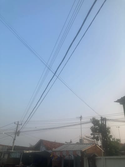The image shows a view of electrical wires crisscrossing a hazy sky above a collection of rooftops in Proyonangg, Indonesia. The wires are a prominent feature, stretching diagonally across the frame and creating a dense network in the middle ground. Below the wires, several buildings with terracotta roofs are visible. Some structures appear to be homes, while others may be shops or small businesses, indicated by the presence of awnings and signs. A utility pole with numerous wires attached is positioned on the left side of the image. Trees are scattered among the buildings, and an antenna is visible on a pole. The overall atmosphere is somewhat subdued due to the haze, which softens the details of the background and creates a muted color palette dominated by blues, grays, and muted earth tones. There are no people clearly visible in the image, and the scene appears to be a typical street or residential area in a town or city.