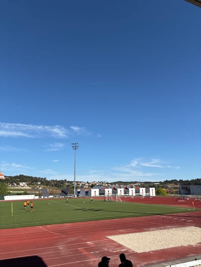 The image shows a partially visible sports complex in Valpaços, Portugal. The sky is mostly clear with a few wispy clouds and a contrail from an aircraft. A tall floodlight pole stands near a running track. The track is red with white lane markers and borders a green field. Several people, dressed in orange and red uniforms, are on the field. White buildings with solar panels are visible in the background, nestled among trees. The edge of a sand pit is also visible.