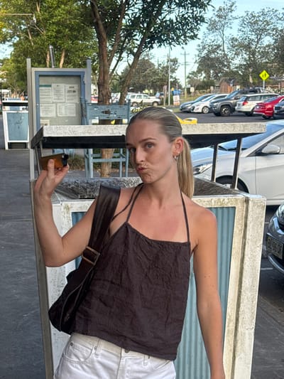 A fair-skinned woman with blonde hair pulled back in a ponytail stands outside, near a road with parked cars. She wears a dark brown tank top with spaghetti straps, white pants, and a dark shoulder bag. She holds a small, brown object in her hand and makes a pouting face at the camera. The background includes trees, street signs, and a concrete structure with a sign.