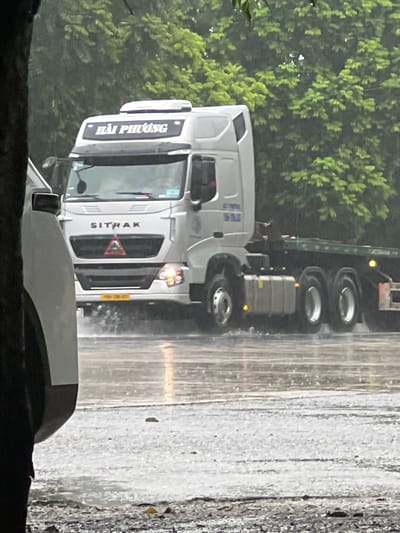 An image captures a white Sitrak truck with "HẢI PHƯƠNG" written on its windshield, driving on a wet road during a downpour, possibly in Hợp Hòa, Vietnam. The truck's headlights are illuminated, and water splashes around its wheels. The road surface is reflective, indicating standing water and heavy rain. In the background, lush green trees line the road, their foliage appearing damp and glossy due to the rain. A portion of a white vehicle is visible on the left side of the frame, partially obstructing the view of the truck. The overall environment suggests a rainy day in a semi-urban or rural setting with abundant vegetation.