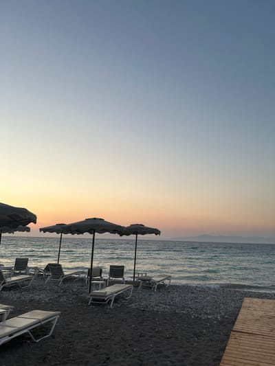 This image captures a serene beach scene in Rhodes, Greece, likely during either sunrise or sunset. The foreground features a dark, pebble-strewn beach with several empty lounge chairs and open umbrellas. A wooden boardwalk runs along the right side of the frame, leading towards the water. The middle ground is dominated by the calm, shimmering sea, with gentle waves lapping at the shore. In the distance, a faint outline of islands or a coastline is visible against the horizon. The sky displays a soft gradient of colors, transitioning from a pale blue at the top to warm hues of orange and yellow near the horizon, indicative of twilight. There are no people or animals present in the scene.