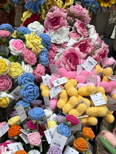 The image displays a dense, close-up perspective of numerous handmade crocheted items, predominantly flowers, arranged in what appears to be a retail or market setting. The foreground and midground are filled with these items, showcasing a broad spectrum of colors.
**Subjects/Objects:**
1. **Crocheted Flowers:** Various styles and sizes are present.
* Large, multi-layered rose-like flowers in shades of pink and fuchsia are clustered in the upper right and central areas.
* Medium-sized rose or ranunculus-style flowers in colors including pink, yellow, white, and blue are distributed throughout the scene, some featuring green leaf-like bases.
* Smaller, individual bloom forms resembling roses or similar flowers are visible in light blue, orange, purple, pink, yellow, and white, typically with a green base.
2. **Other Crocheted Forms:**
* Numerous elongated, yellow crocheted objects, suggestive of peanuts or small stuffed pods, are grouped in the mid-right section of the frame.
* Pink crocheted heart shapes are interspersed among the yellow objects and other floral displays.
**Details:**
Each item or small cluster of items has a small white rectangular tag attached. Some tags feature barcodes and alphanumeric sequences, while others display the text "DDD" accompanied by three star symbols. The items are arranged within what appear to be woven baskets or display containers. The background is softly blurred, showing additional items, possibly more crocheted goods or craft supplies, and hints of a structured display.
**People/Actions/Interactions:**
No people are visible in the image. No specific actions or interactions are depicted; the scene is static.
**Environment:**
The items are arranged in a manner consistent with a commercial display, such as a craft fair stall, a market vendor's display, or a section within a retail store specializing in handmade goods.
**Location:**
The image does not provide any visual cues, such as specific signage, unique architectural features, or identifiable landmarks, to pinpoint its precise location within the city of Sint-Niklaas, Belgium.
