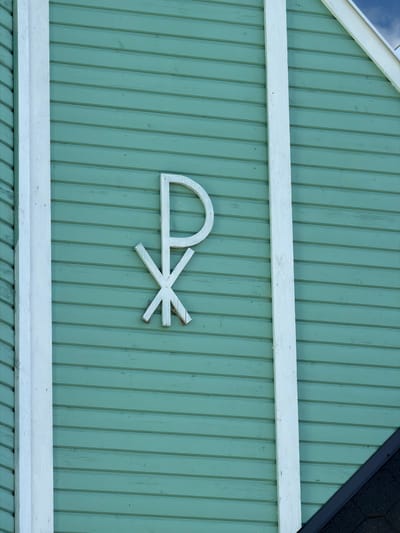A close-up, eye-level exterior shot shows a light blue-green wooden clapboard wall of a building. A white wooden Chi Rho symbol is affixed to the wall, off-center to the right. The symbol is composed of a Greek chi (Χ) superimposed on a Greek rho (Ρ). The left edge of the building features a vertical white wooden trim. A dark, shingled roofline is visible in the lower right corner of the frame, intersecting the wall at an angle. The image is taken on a bright, possibly sunny day, with a patch of sky visible in the upper right corner. The location is the Church of the Assumption of the Most Holy Mother of God in Läskelä, Russia.